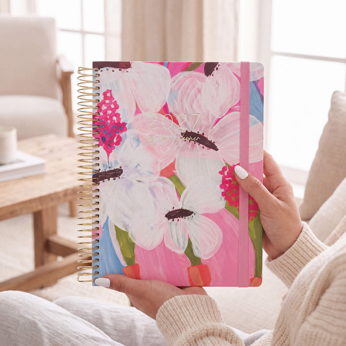 2027 Signature Planner in Petals held by a woman in a cream knit sweater, gold coil and pink elastic closure.