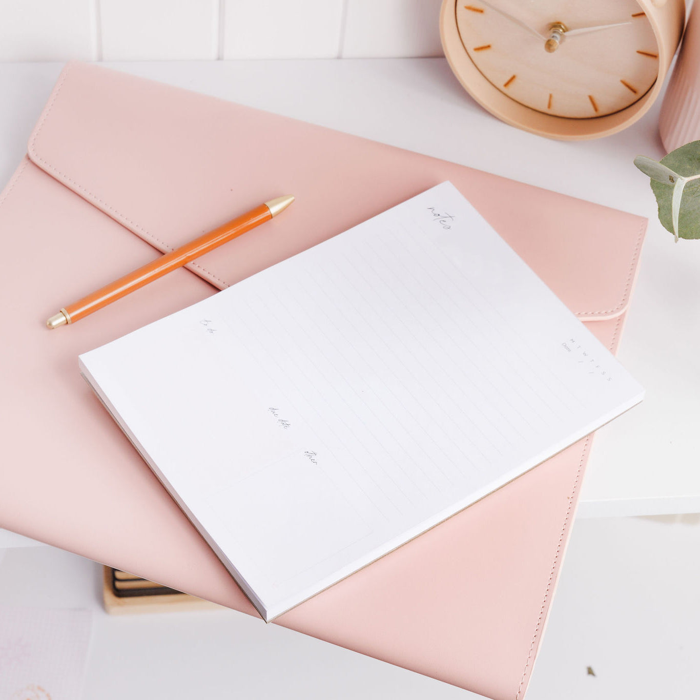 A5 notepad with sections for notes and a to-do list, sitting on a folio at a side angle on a desk with a pen beside it.