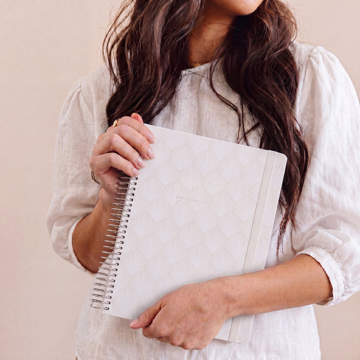 2027 Signature Planner in Shells held by a woman in a white linen shirt, silver coil and cream elastic closure.