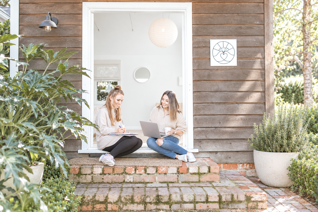 Two teachers sitting on a sunny doorstep, one working on a laptop with a coffee in hand, the other writing in a teacher planner