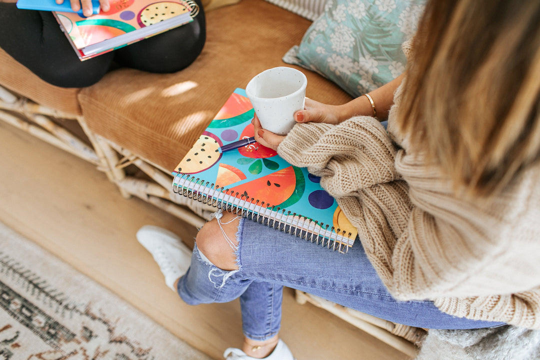 Overhead shot of a teacher sitting on the floor with a mug of tea and a brightly patterned spiral teacher planner resting on her lap
