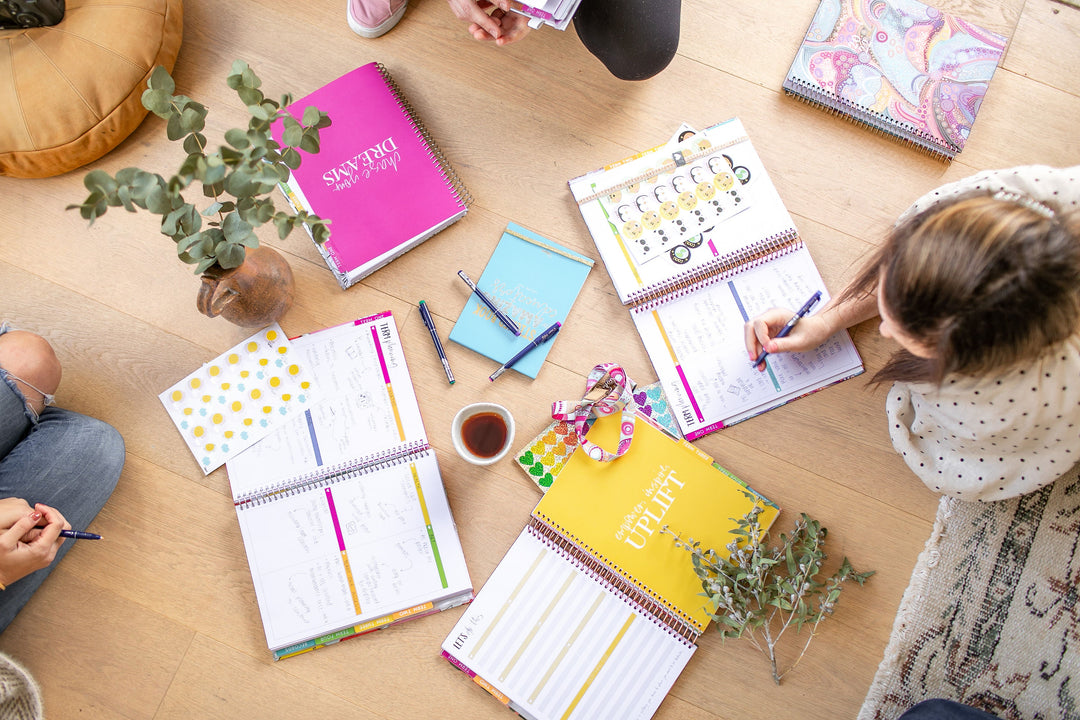 Overhead view of a timber table with several open Mrs Edgar planners and notepads being written in, alongside pens, a potted eucalyptus and a cup of tea