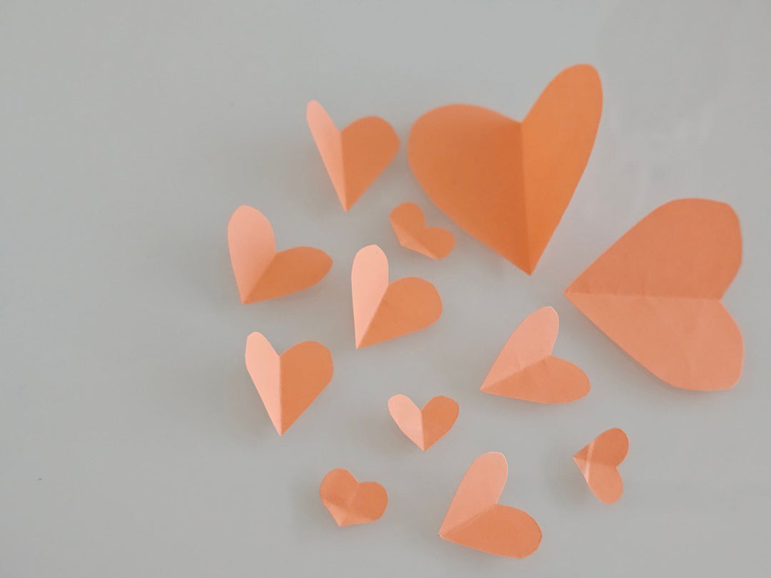 Folded paper hearts cut from coral and peach paper, scattered across a soft grey surface