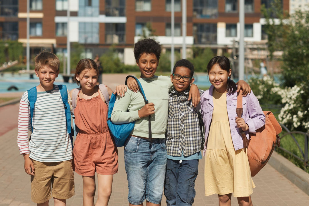 Five smiling school children with backpacks standing outdoors with their arms around each other's shoulders