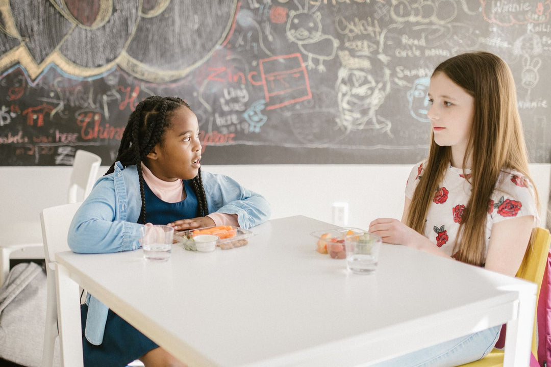 Two primary students sitting across a classroom table with lunch containers, one talking while the other listens