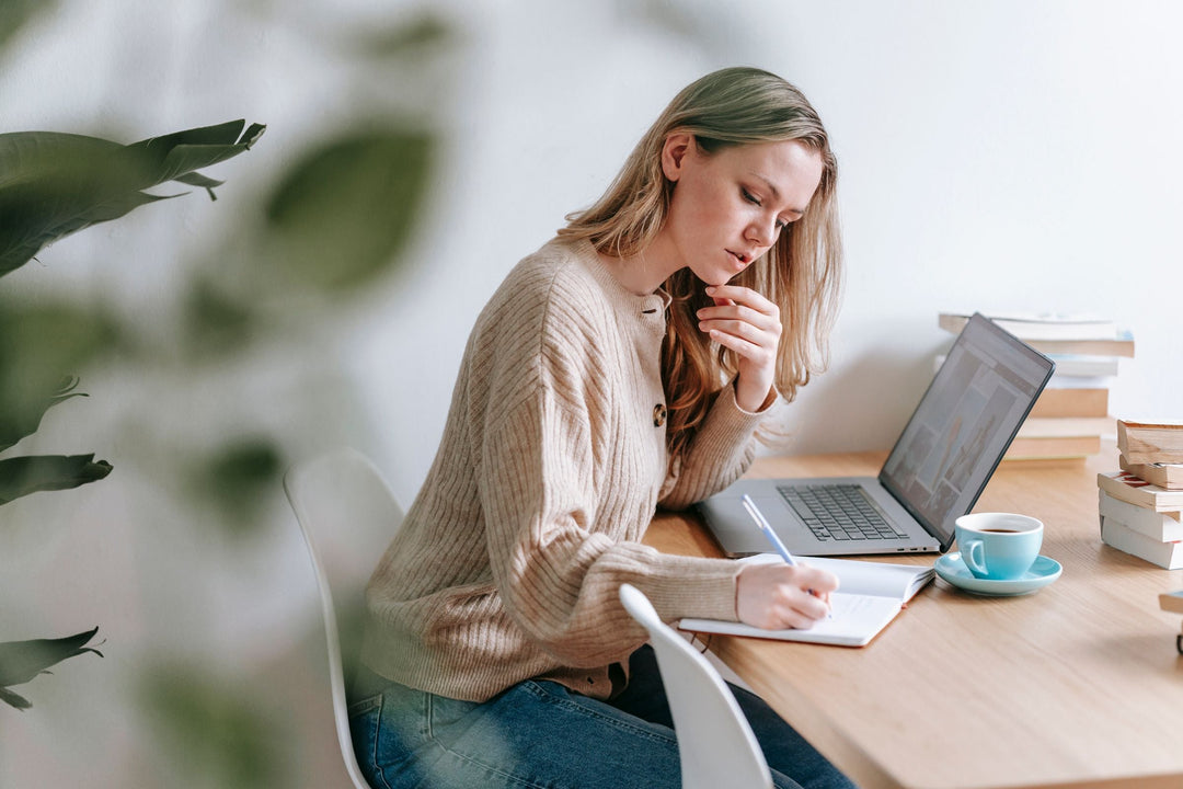 A primary teacher sitting at a wooden desk working on an open laptop, taking notes in a notebook with a cup of coffee beside her