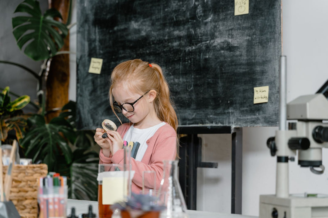 A girl in glasses studying a small object through a magnifying glass beside a microscope in a classroom