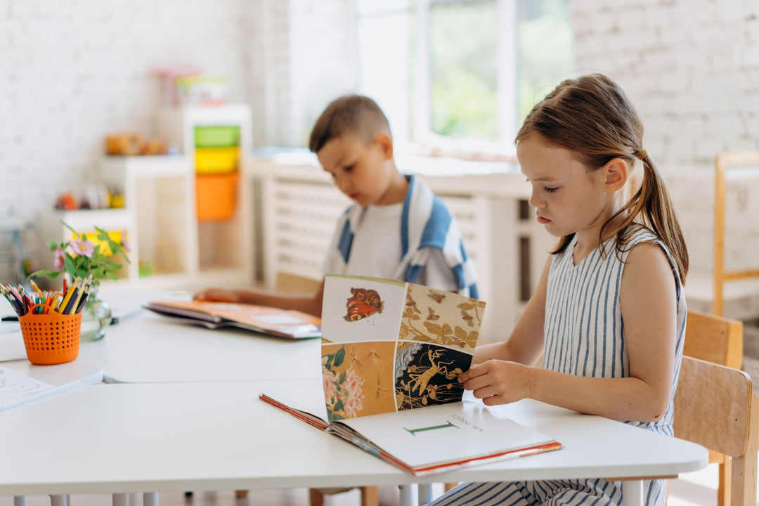 Two primary school children sitting at white desks turning the pages of open illustrated books in a bright classroom with pots of coloured pencils
