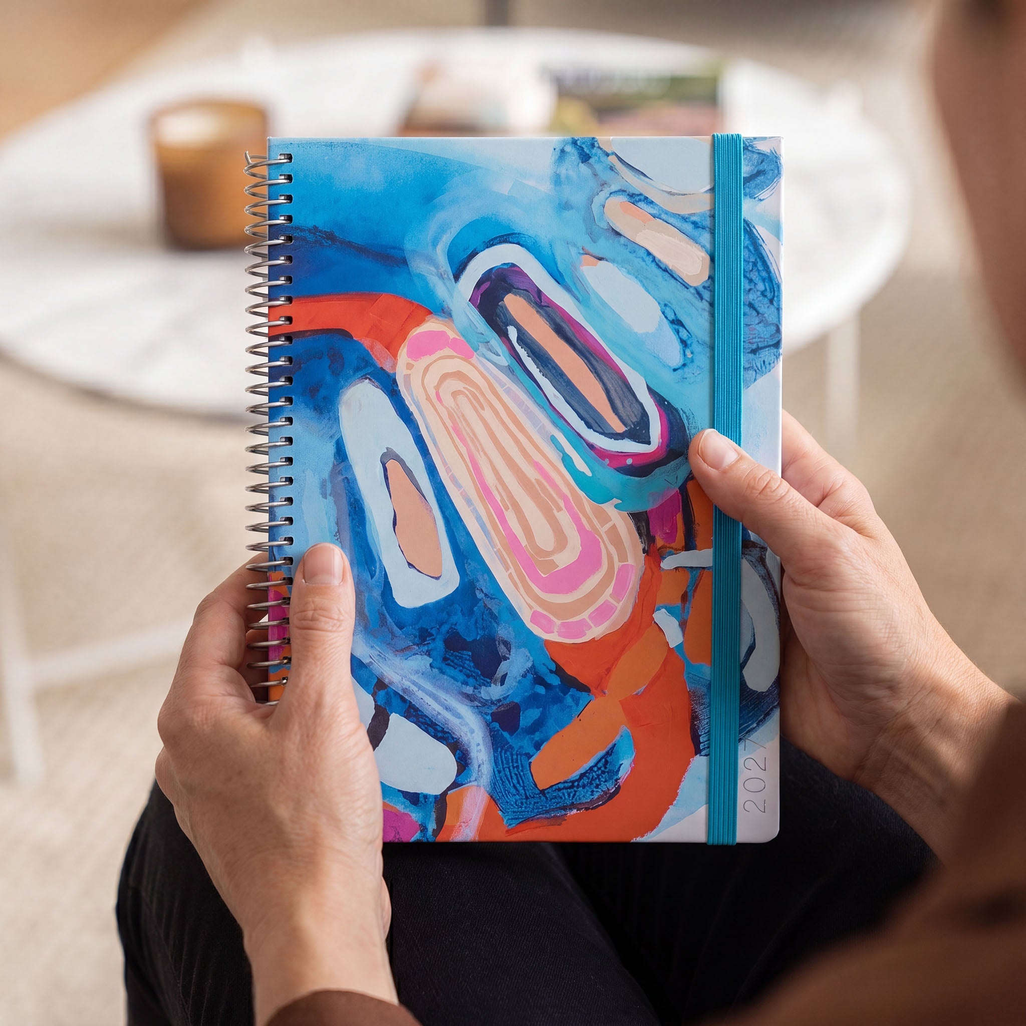 Hands holding a 2027 slimline spiral planner with bold abstract blue and orange artwork and teal elastic closure.