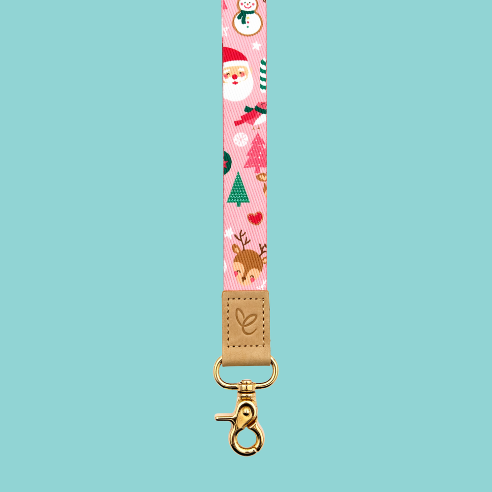 Pink festive lanyard with holiday characters including reindeer and Santa, finished with a tan leather logo patch and gold hardware on a blue background.