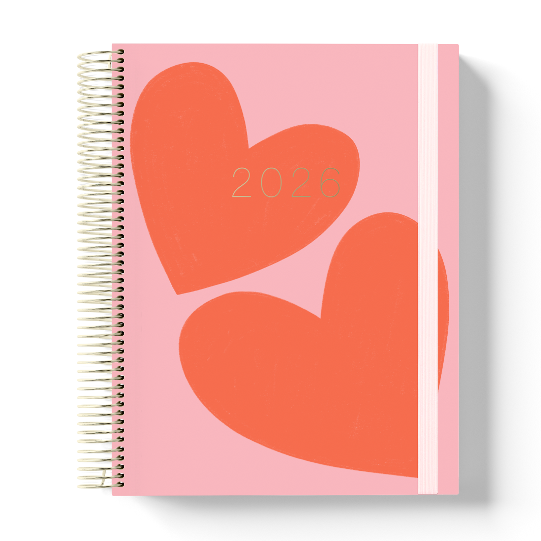 2026 teacher planner with a light pink cover featuring two large red-orange hearts, gold coil binding, and white elastic strap.