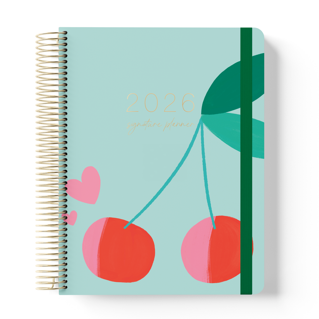 2026 signature planner with cherry and heart illustrations on a pale mint green cover, spiral-bound with a green elastic strap.