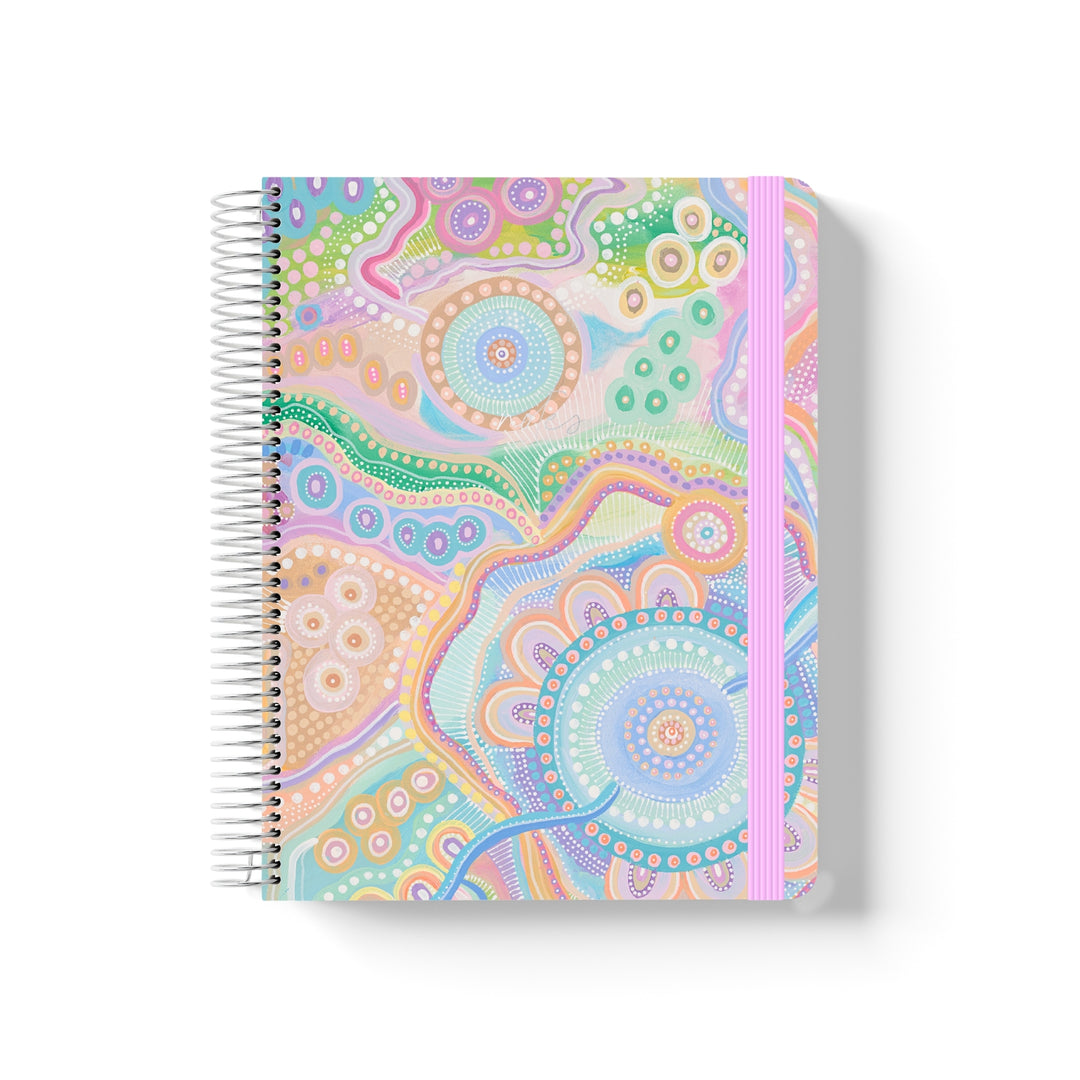 Coiled notebook with pastel Indigenous artwork by Holly Sanders, pink elastic band, and intricate circular and dot motifs.