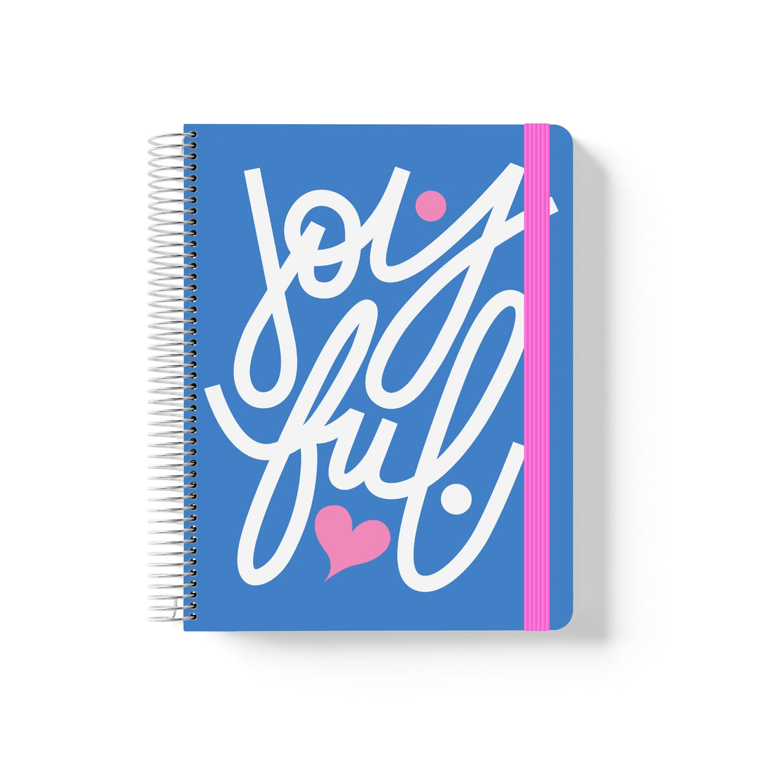 Blue coiled notebook with pink elastic band and bold white script reading “joyful” with a heart and dot accents.