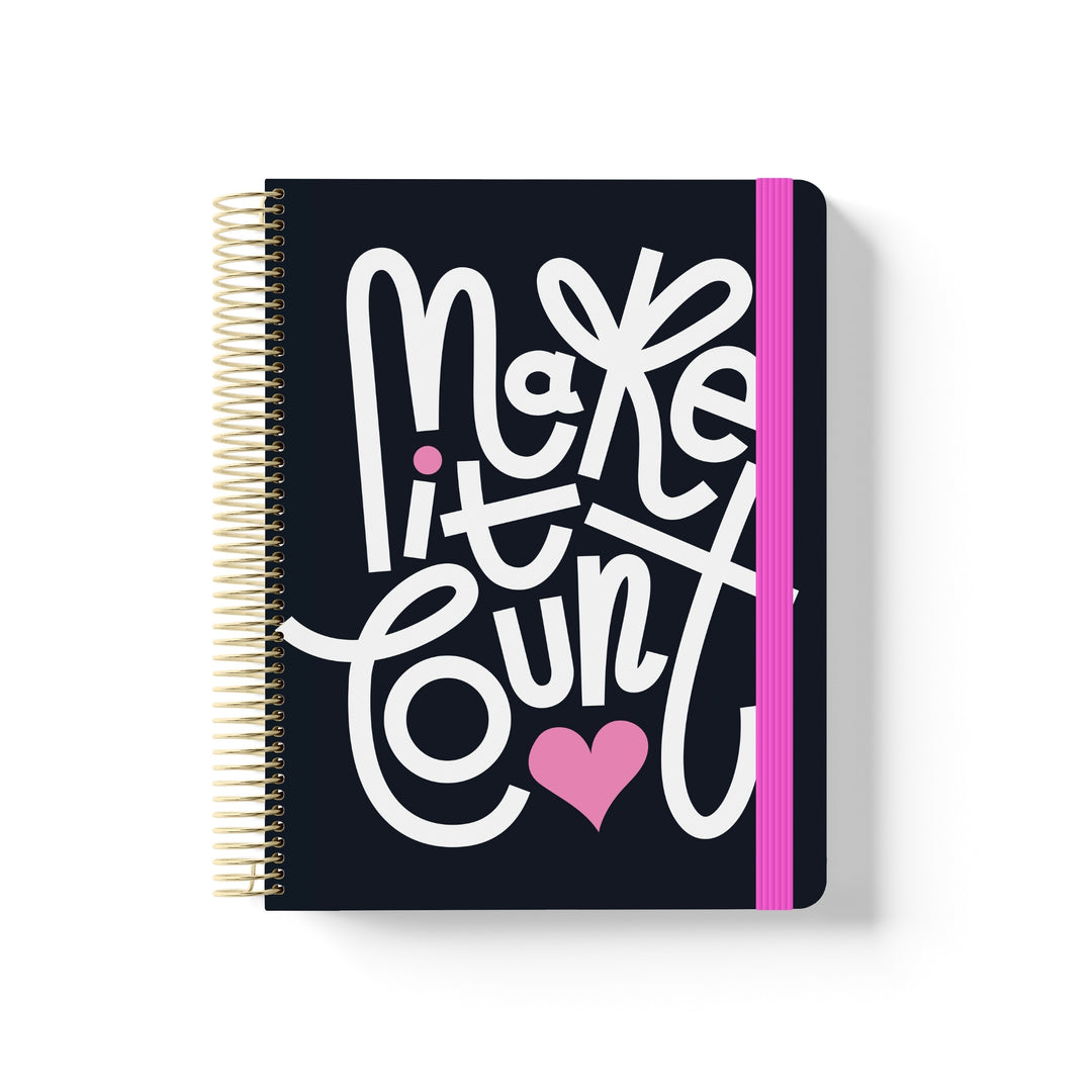 Black coiled notebook with bold white hand-lettered text “Make it count,” pink heart and dots, gold spiral, and magenta elastic band.