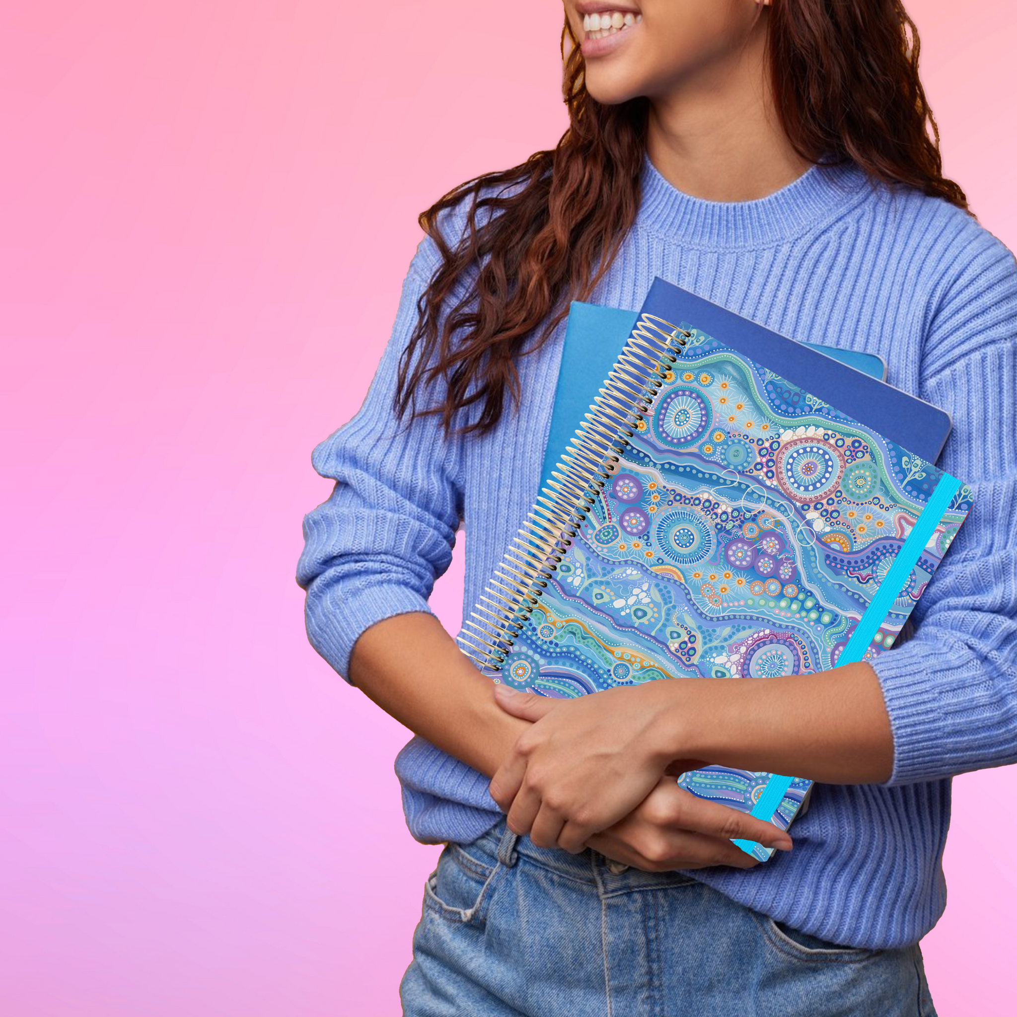 Smiling woman in a blue sweater holds the 2026 teacher planner featuring blue Aboriginal art design on the cover.