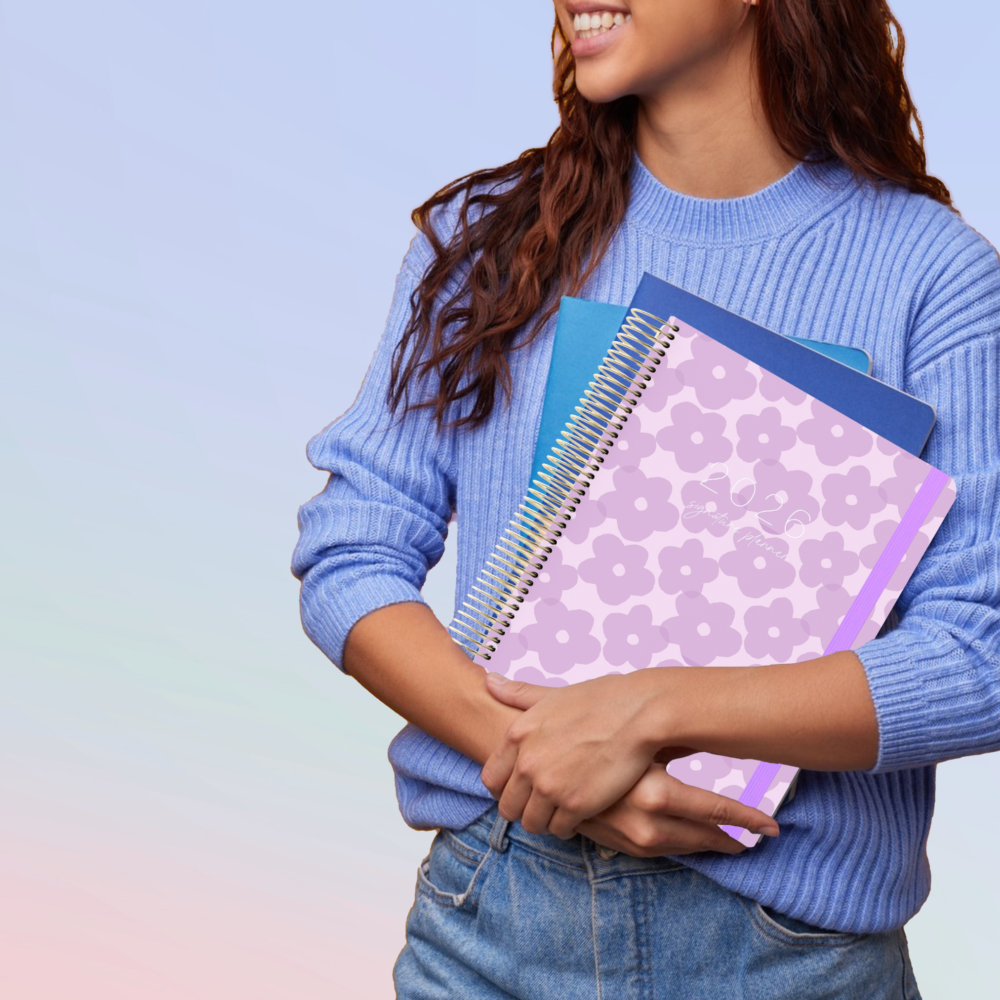 Smiling woman in blue sweater holding a lilac floral planner with gold spiral and purple elastic, alongside two blue notebooks.