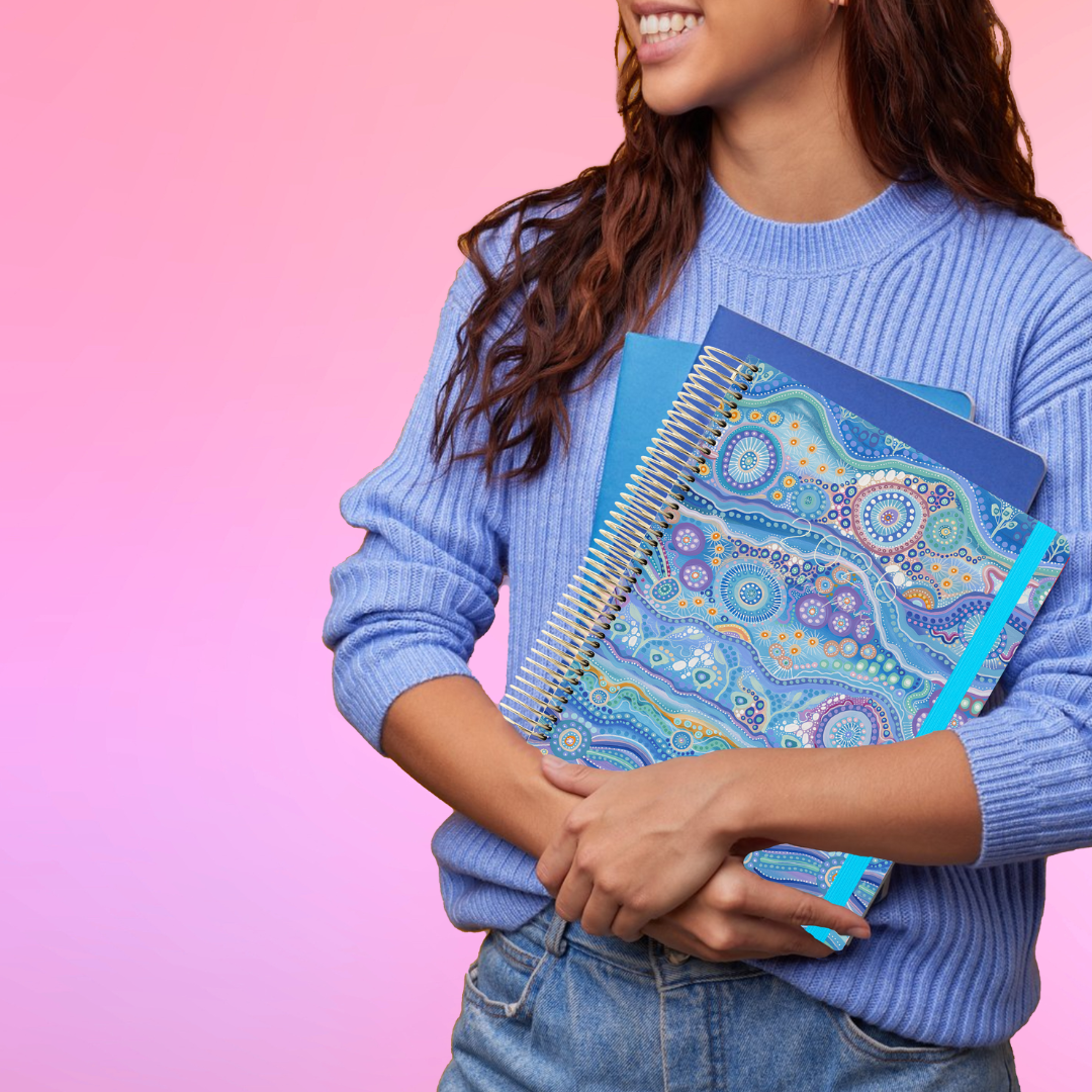Smiling woman in a blue sweater holds the 2026 teacher planner with blue Aboriginal art design on the cover.