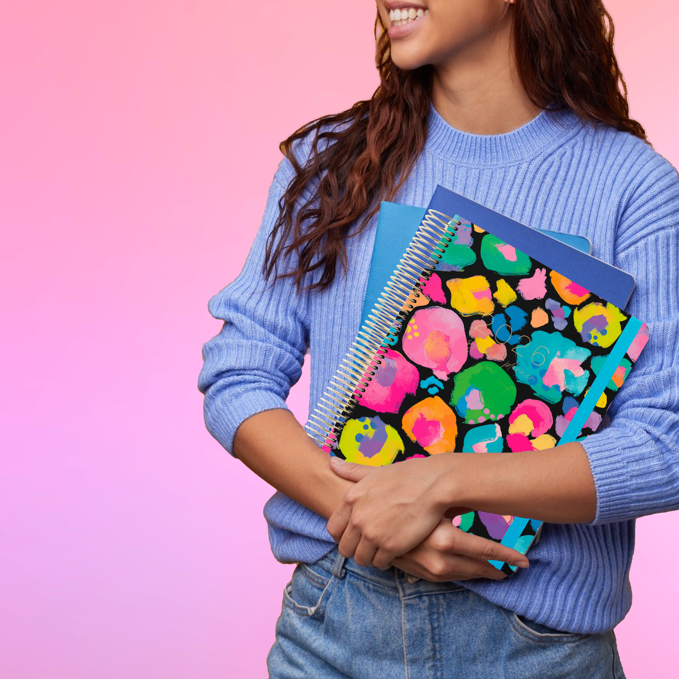 Smiling woman in a blue sweater holds the 2026 teacher planner with bright colourful splotches on the cover.