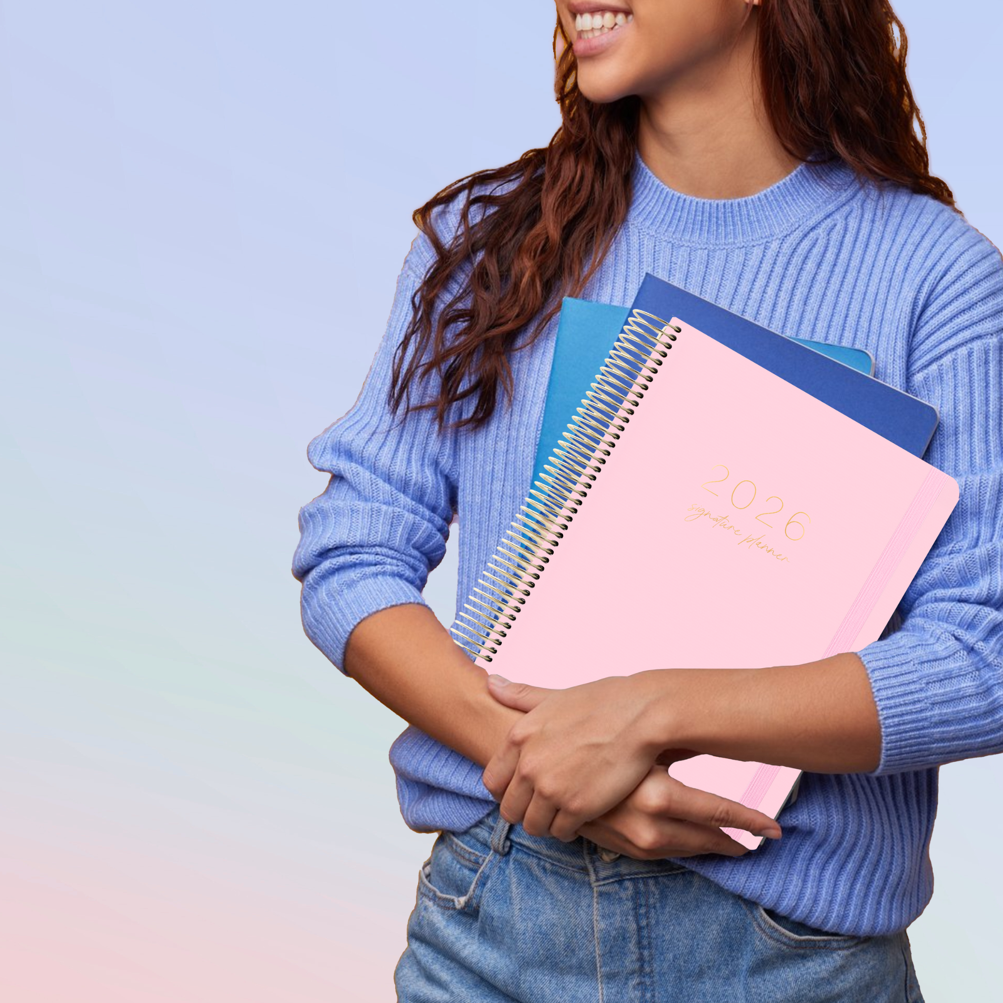 Woman in blue sweater holding a blush pink 2026 teacher planner with gold coil and elastic strap, paired with blue notebooks.