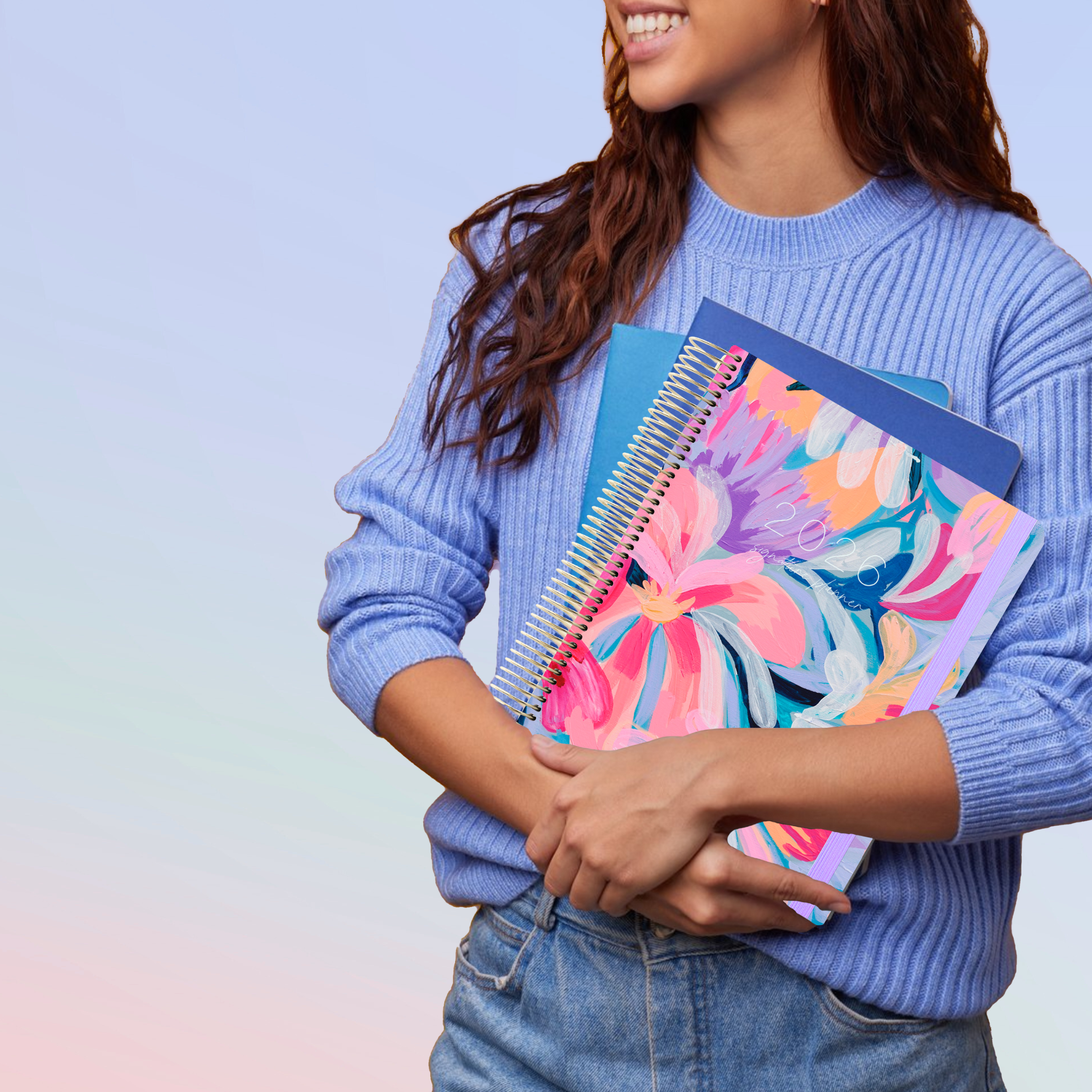 Smiling woman in blue sweater holding a vibrant floral 2026 planner with gold spiral and lilac elastic, alongside blue notebooks.
