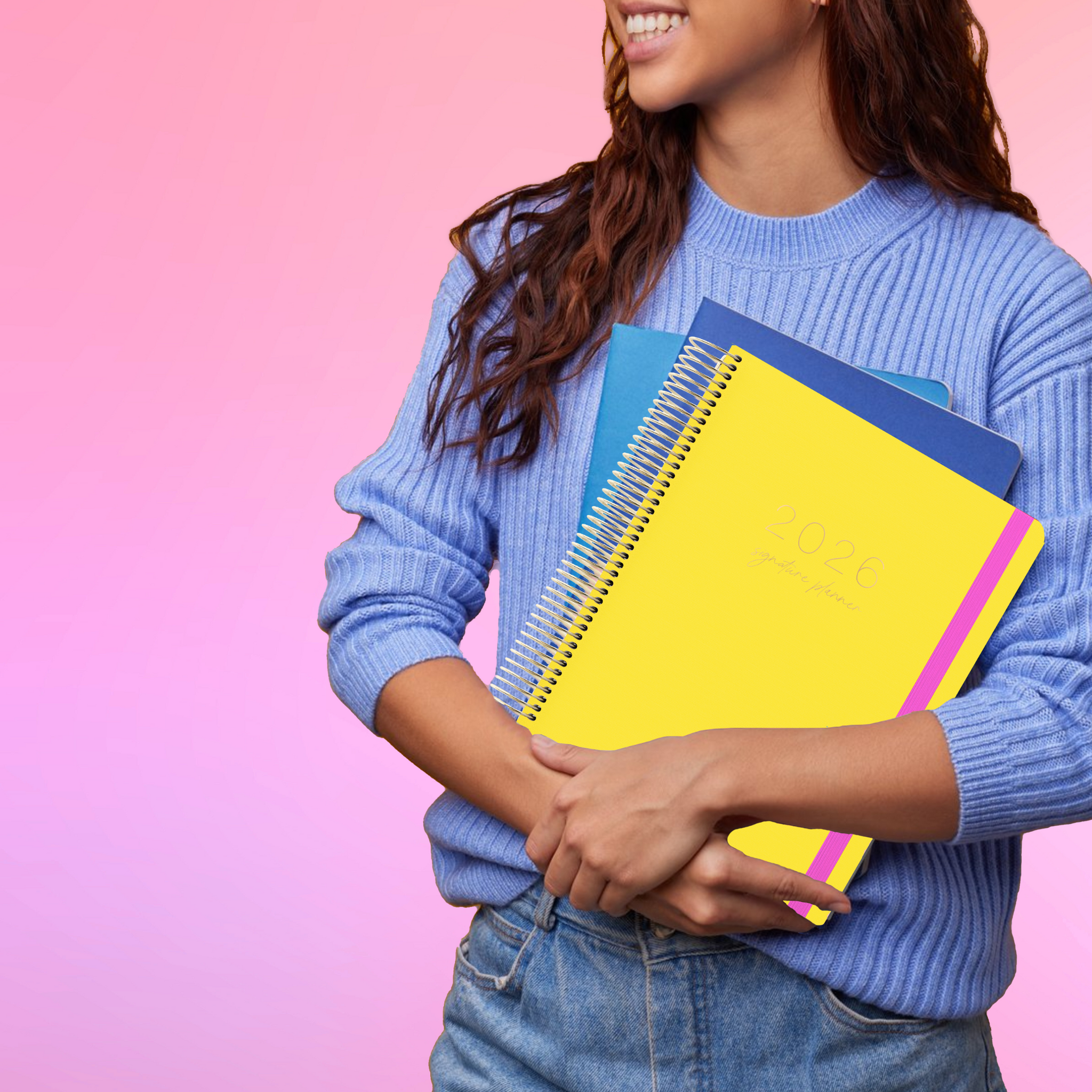Smiling woman in a blue sweater holding a 2026 yellow spiral planner against a pink gradient backdrop.