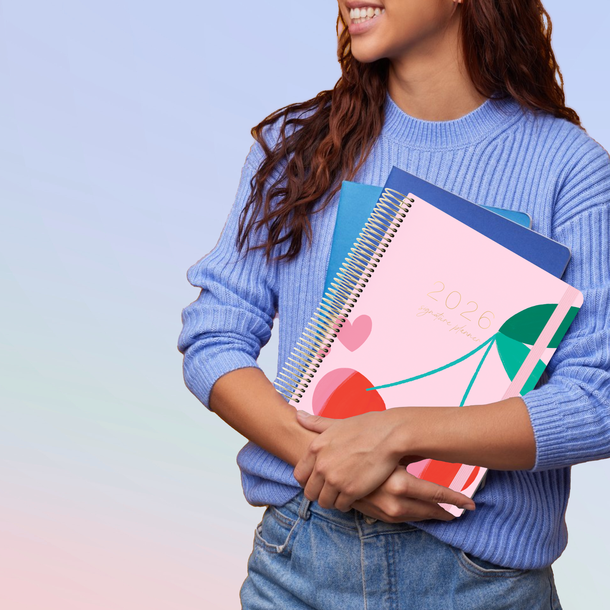 Woman in blue sweater holding 2026 pink cherry planner with gold coil and dark green elastic strap.
