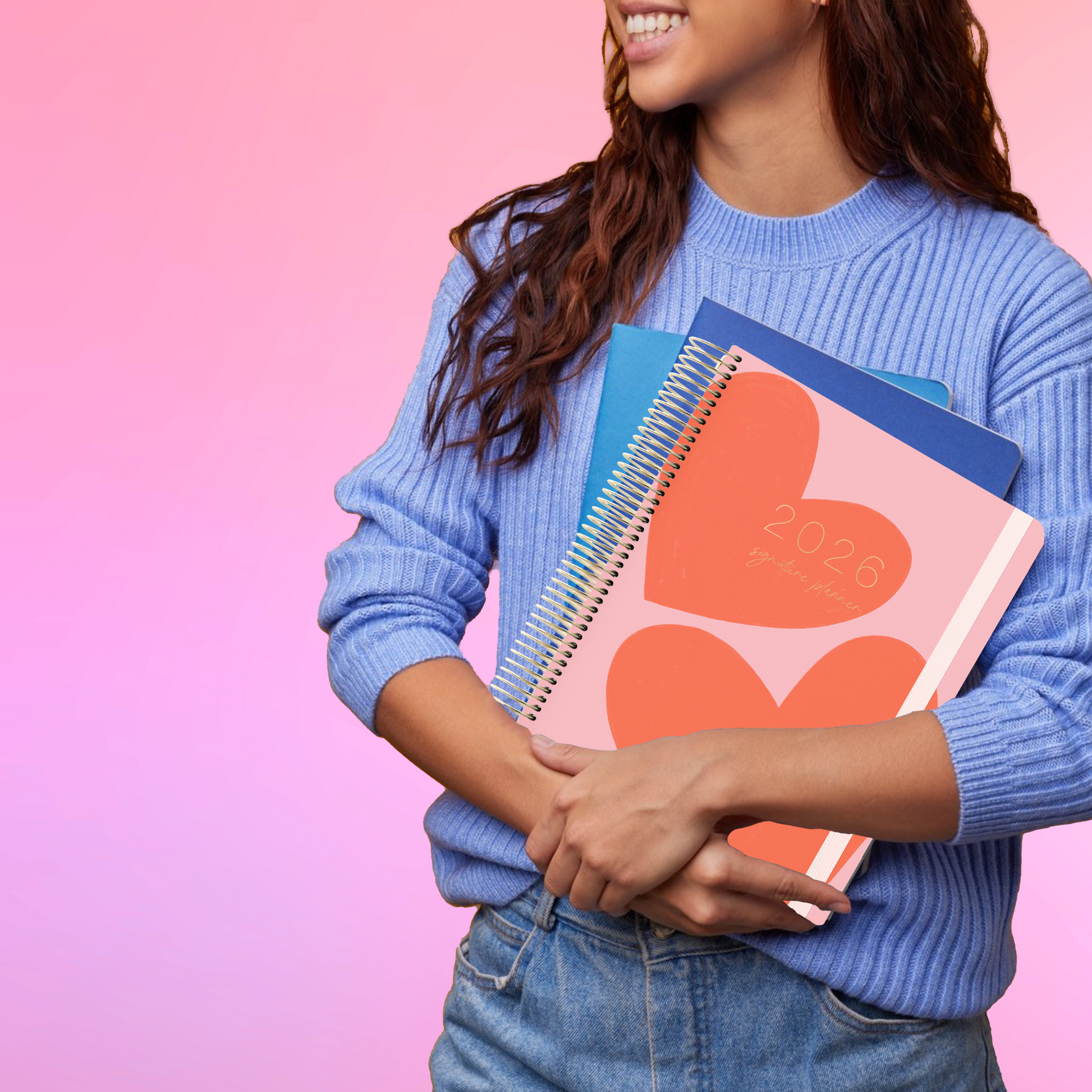 Smiling woman in a blue sweater holds the 2026 teacher planner with a 2 red hearts on a pink cover.