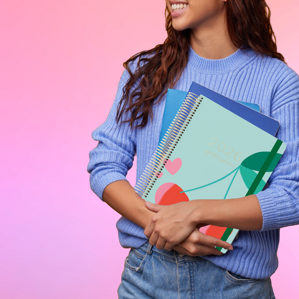 Smiling woman in a blue sweater holds the 2026 teacher planner with a modern cherry and leaf design on the cover.