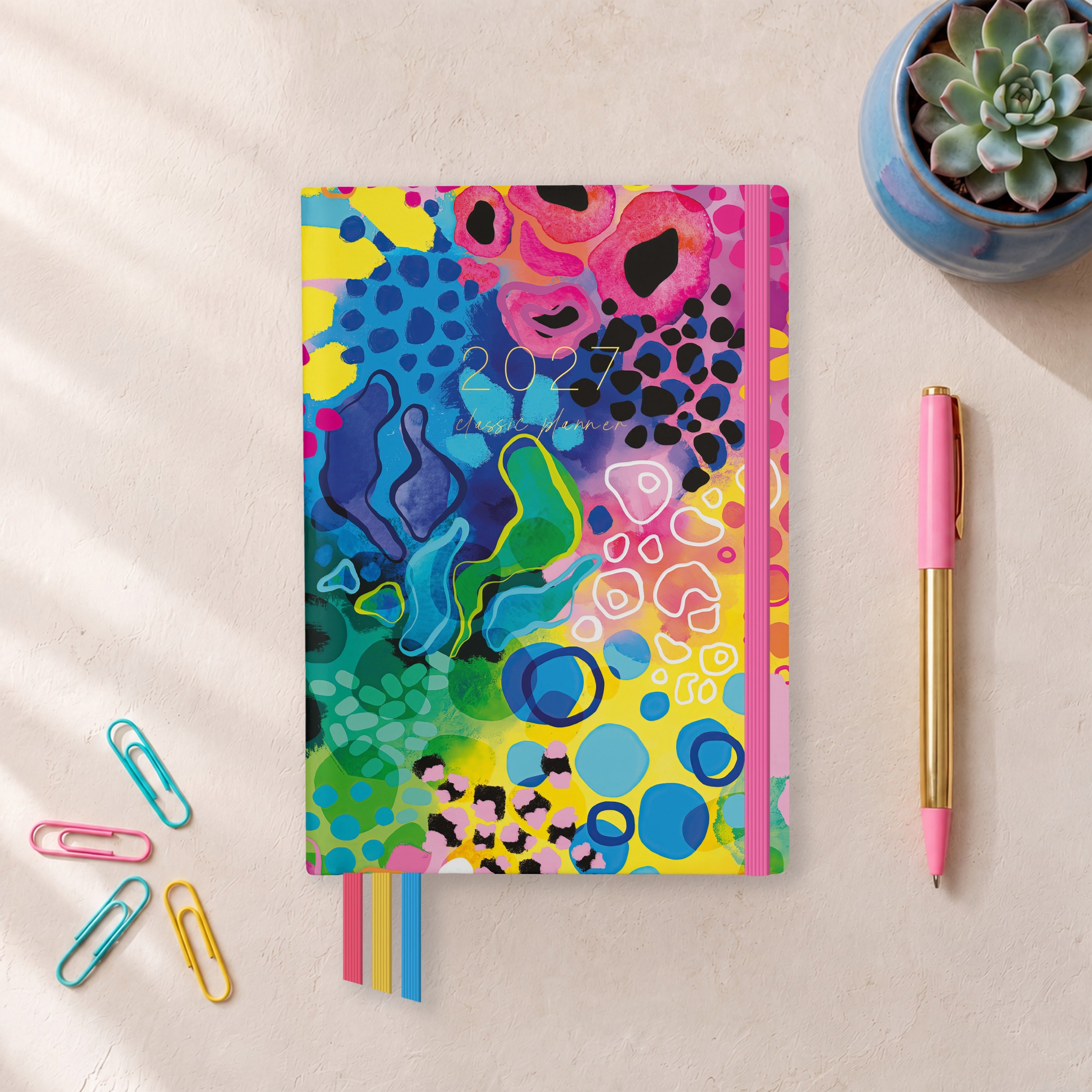 2027 Boom Classic Weekly Teacher Planner flat lay with bold colourful abstract cover, styled with a succulent, pink pen and paper clips