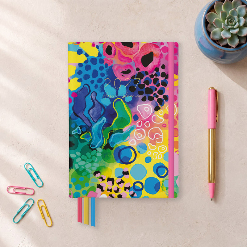 2027 Boom Classic Weekly Teacher Planner flat lay with bold colourful abstract cover, styled with a succulent, pink pen and paper clips