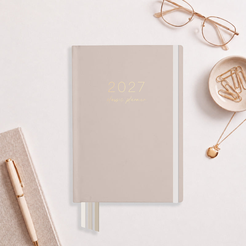 2027 Latte Classic Weekly Teacher Planner flat lay with warm taupe cover and gold foil, styled with gold glasses and an apple pendant