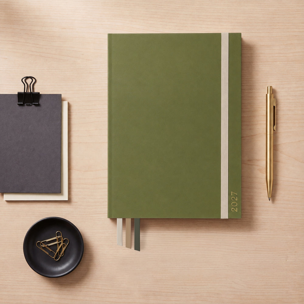 2027 Moss Slimline Classic Teacher Planner flat lay with olive green cover and gold 2027 foil, styled with a brass pen and notepads