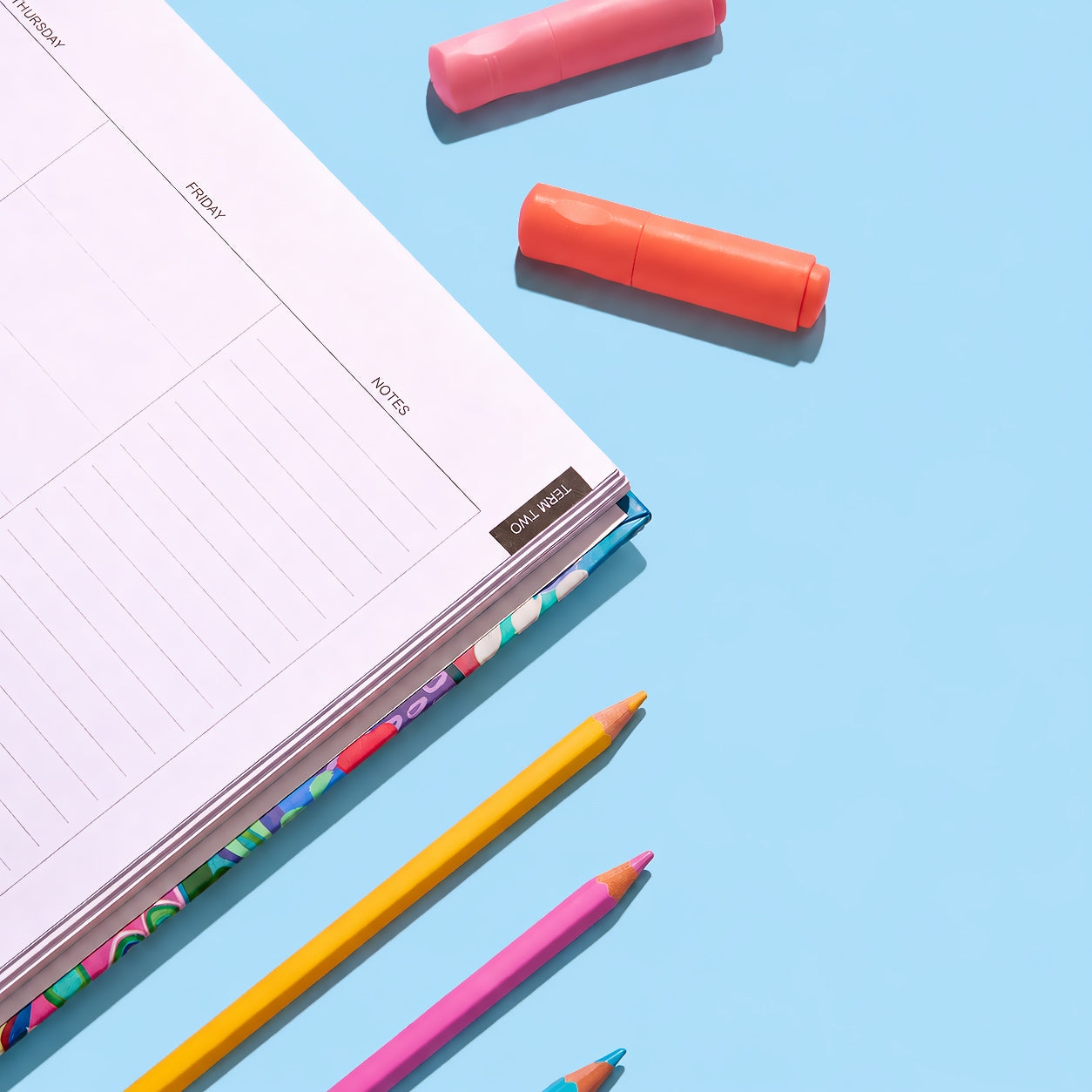 Close-up of the 2027 Classic Slimline Teacher Planner weekly page with Term Two tab and colourful cover edge, styled with pencils and highlighters on a blue background