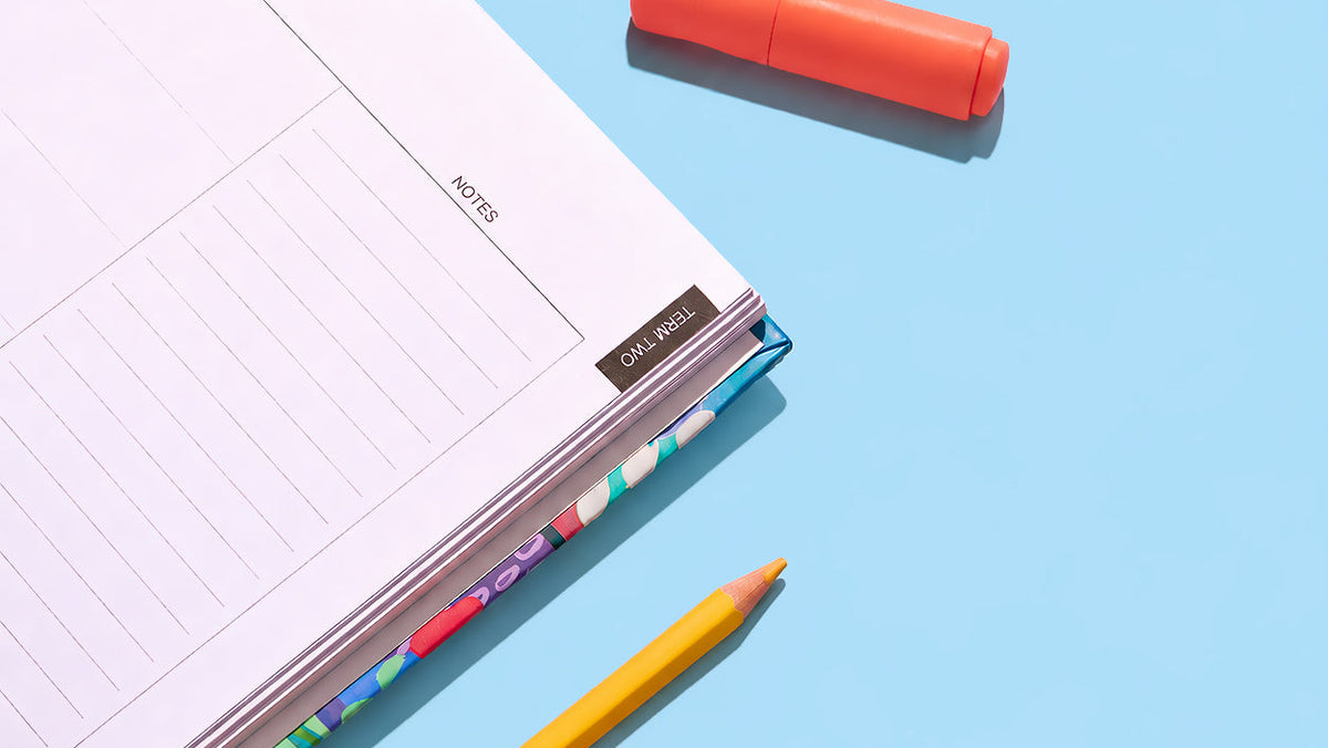 Close-up of the 2027 Classic Slimline Teacher Planner weekly page with Term Two tab and colourful cover edge, styled with pencils and highlighters on a blue background
