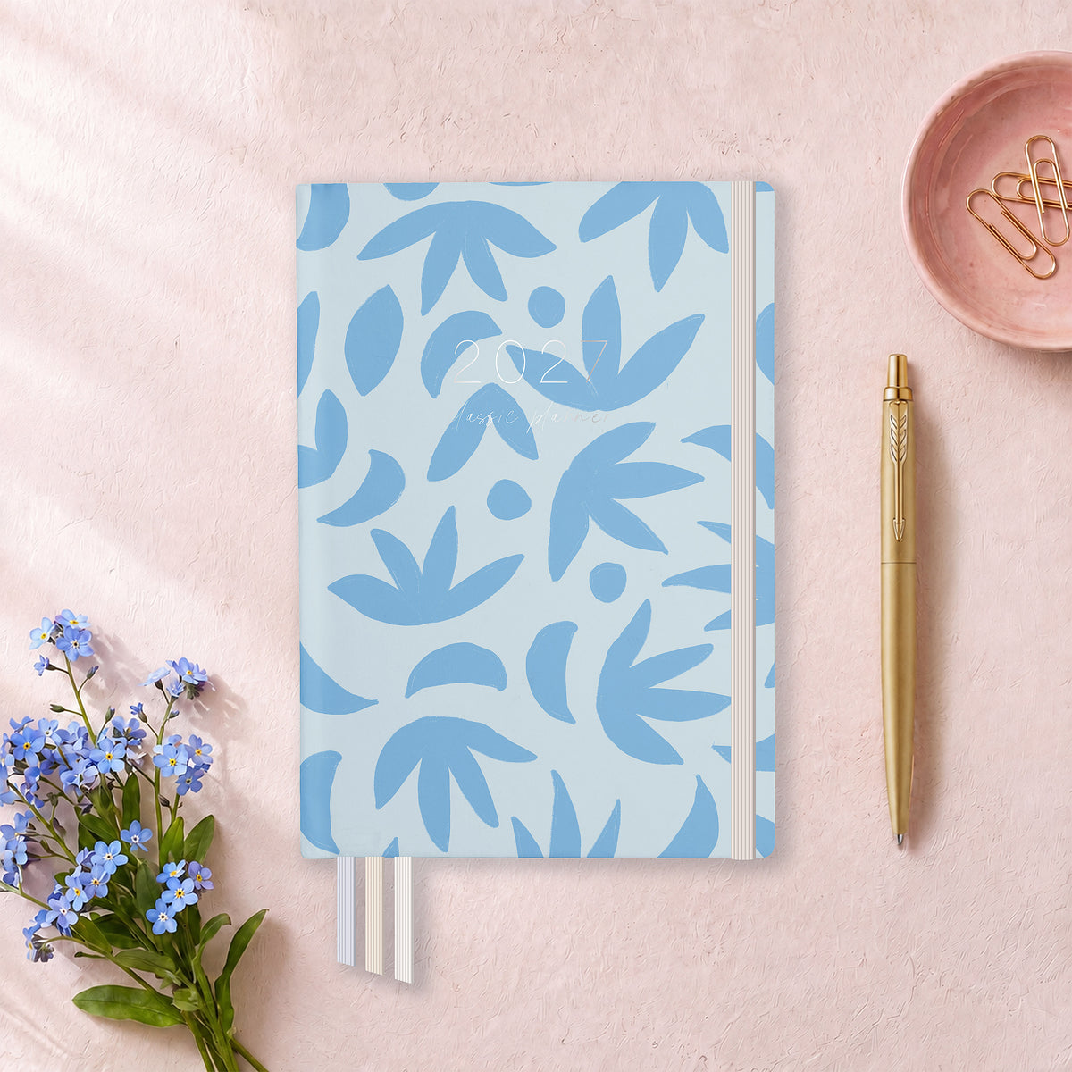 2027 Sunburst Sky Classic Weekly Teacher Planner flat lay with blue leaf pattern cover, styled with forget-me-nots and a gold pen on pink