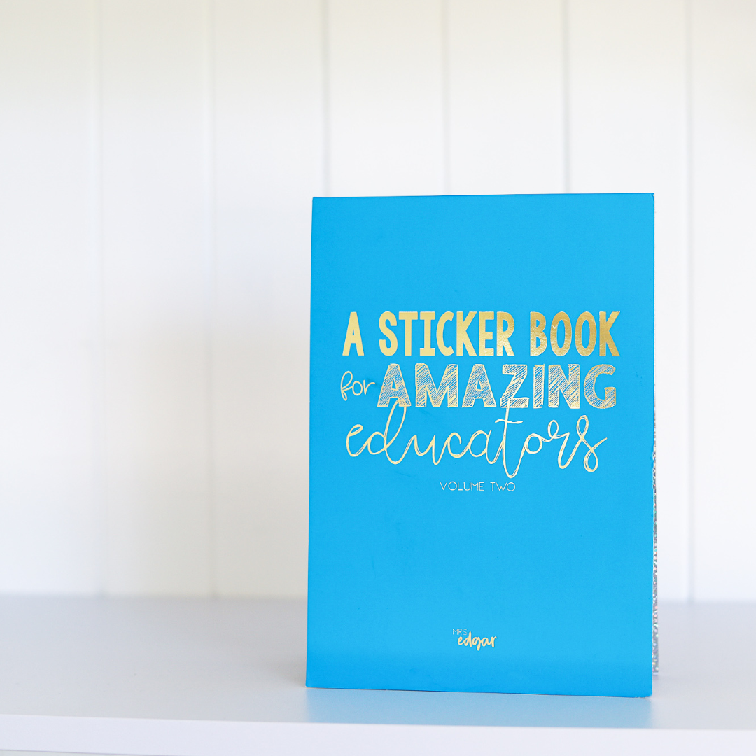 Blue A5 sticker book standing upright on a desk, with gold-foiled text 'A Sticker Book for Amazing Educators Volume 2' and 'Mrs Edgar' at the bottom, featuring a gold elastic enclosure.