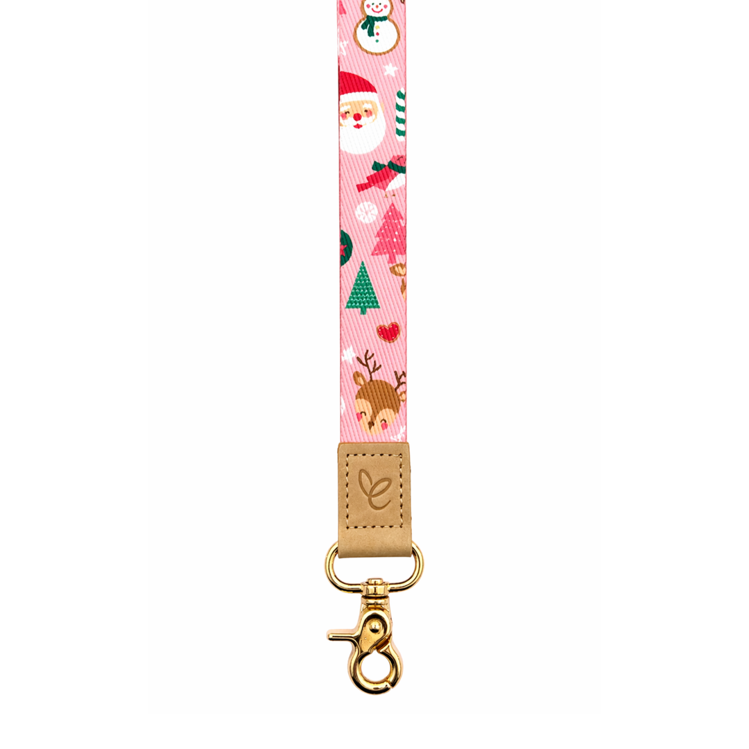 Pink Christmas-themed fabric lanyard with reindeer, Santa and snowman print, featuring a tan leather logo tab and gold clasp on a clean white background.