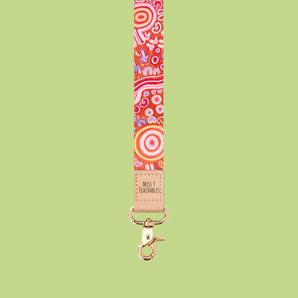Red and pink Indigenous-inspired patterned lanyard with tan leather “Miss T Teachable” logo detail and gold clasp displayed against a soft green background.
