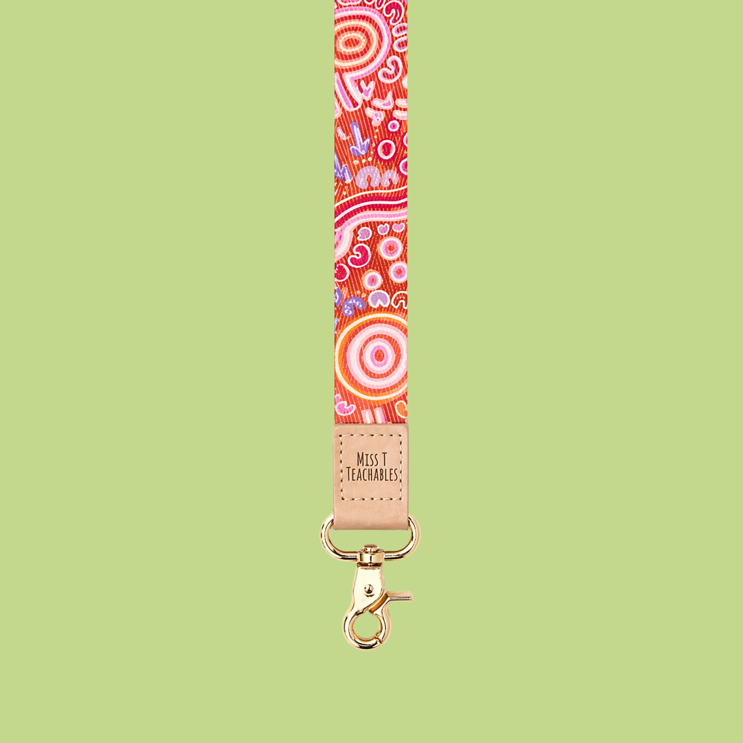 Red and pink Indigenous-inspired patterned lanyard with tan leather “Miss T Teachable” logo detail and gold clasp displayed against a soft green background.