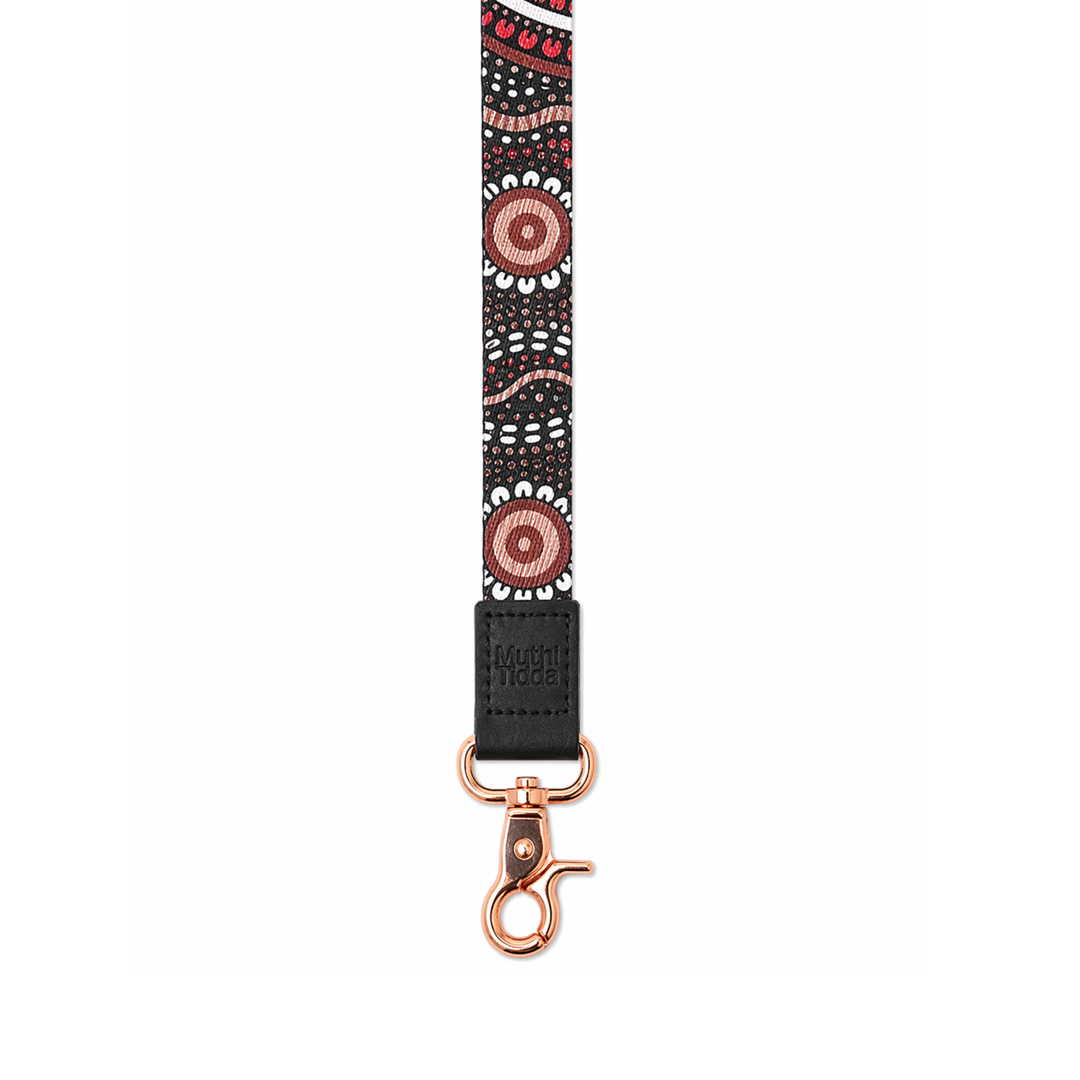 Black patterned fabric lanyard with Indigenous-inspired dot artwork, black leather logo patch, and rose gold clasp on a clean light grey background.