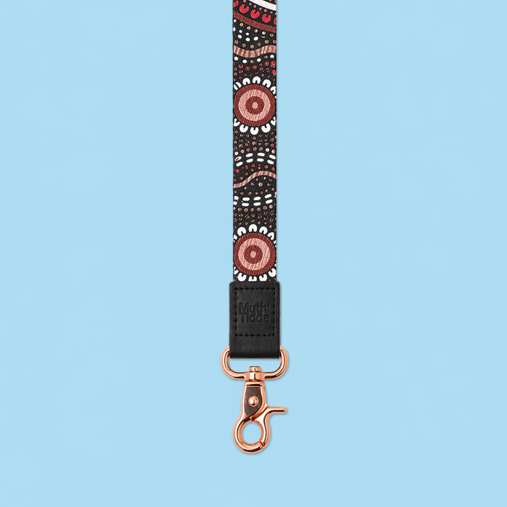 Black fabric lanyard with Indigenous-inspired dot design, black leather logo detail, and rose gold clasp displayed against a soft blue background.