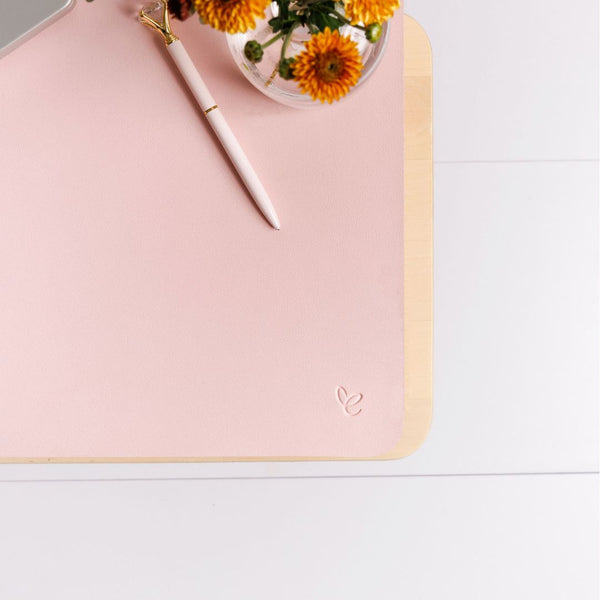 A blush vegan leather desk mat with a small heart embossing, a pen, and a vase of flowers on top, placed on a white desk surface.