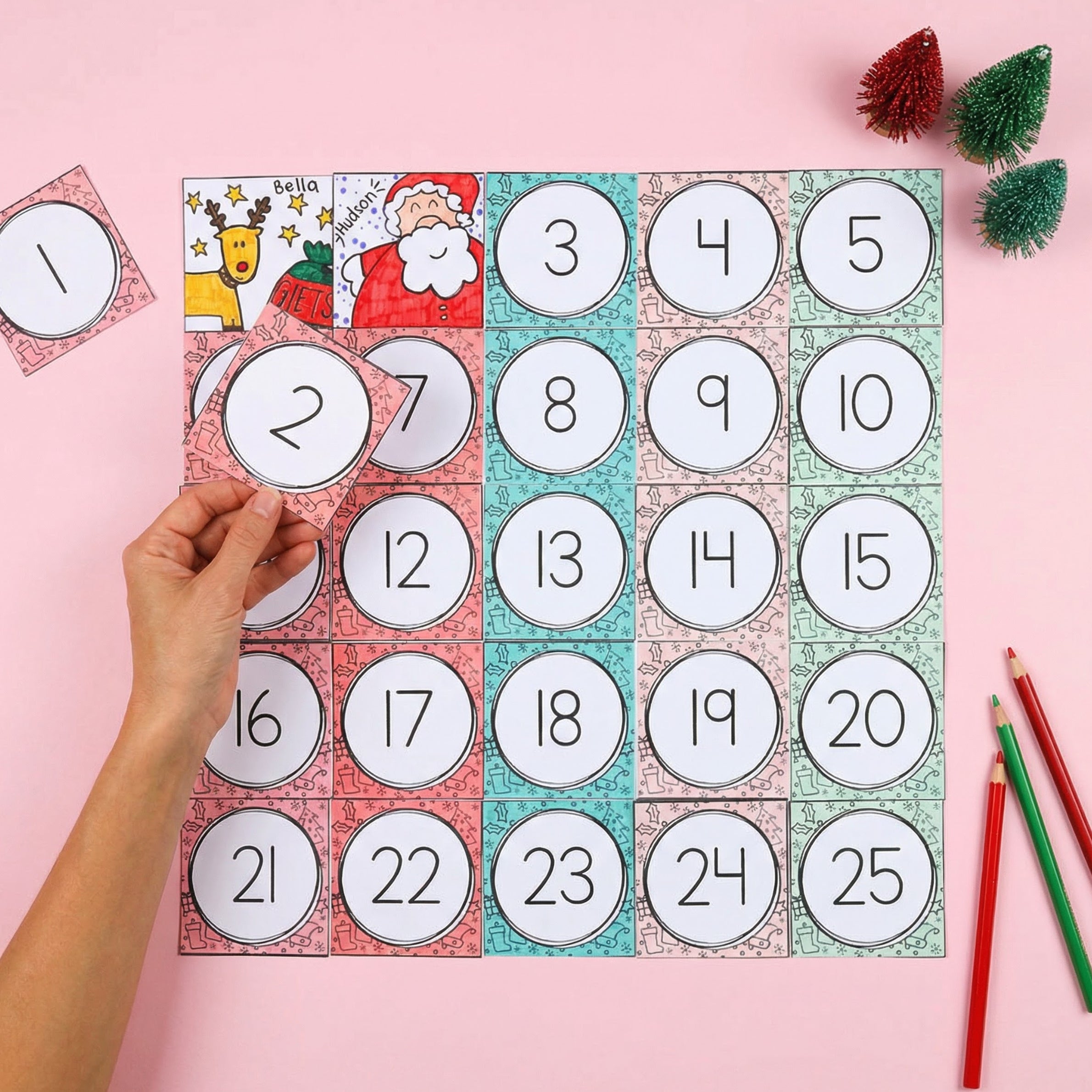 Printable Christmas advent calendar activity with numbered squares from 1–25 and removable number cards placed on top. A hand holds one of the cards while small coloured pencils and mini Christmas trees decorate the pink background.