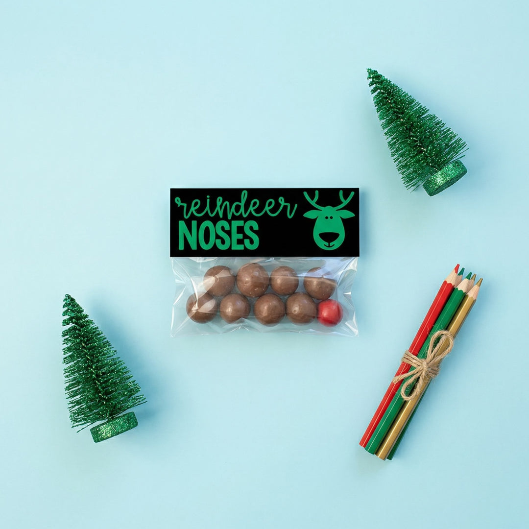 Printable “Reindeer Noses” bag topper attached to a small treat bag filled with chocolate balls and one red candy, styled on a blue background with mini Christmas trees and coloured pencils.