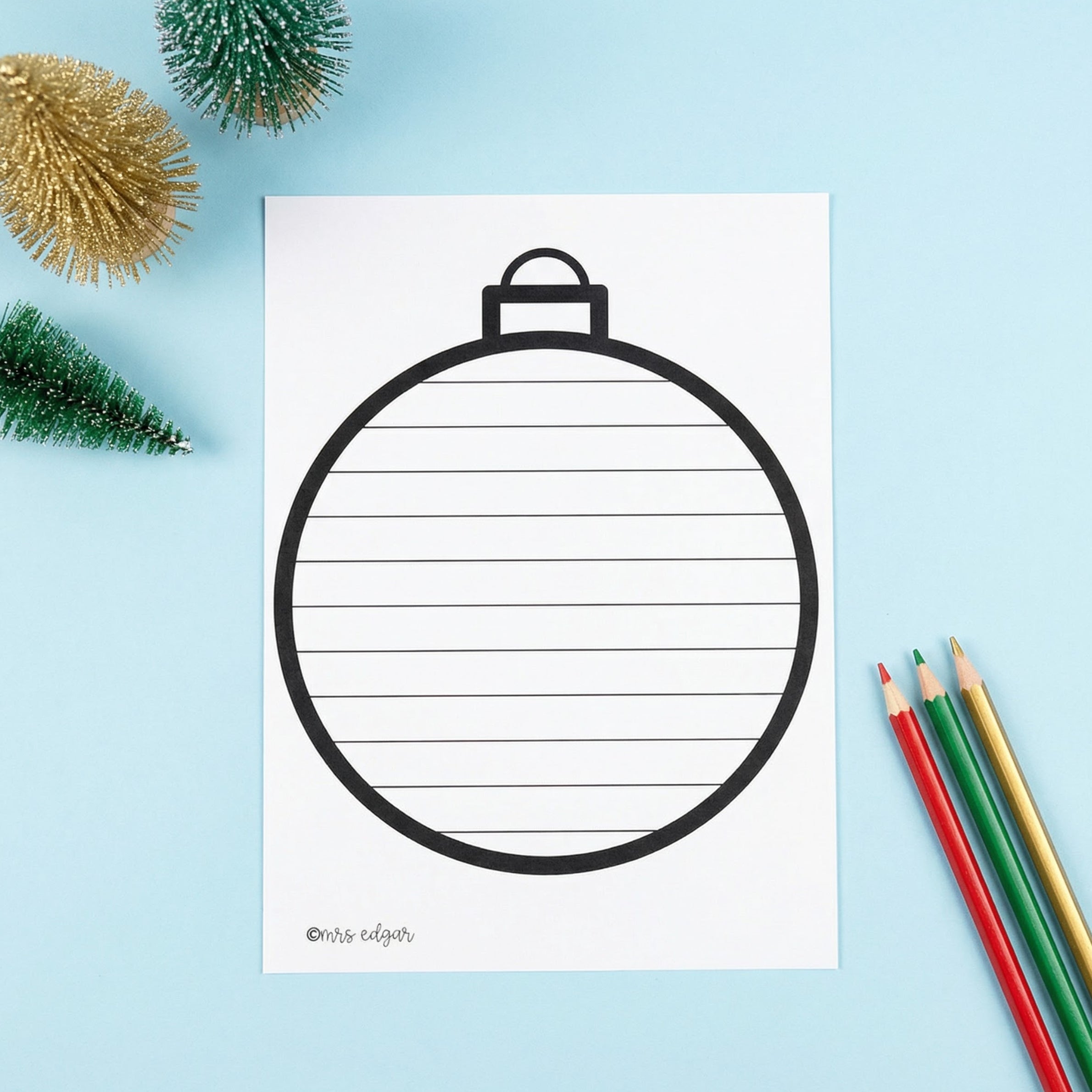 Printable Christmas bauble writing template with lines inside the ornament shape, displayed on a blue background with small Christmas trees and coloured pencils.
