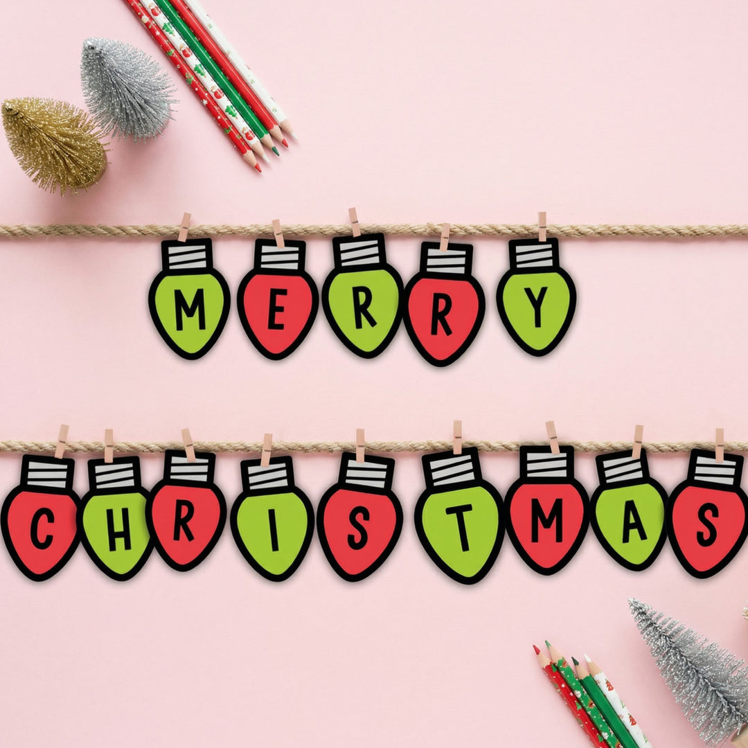 Printable Christmas bunting featuring lightbulb shapes that spell “Merry Christmas,” clipped onto string with small pegs. The display is styled on a pink background with coloured pencils and small decorative Christmas trees.