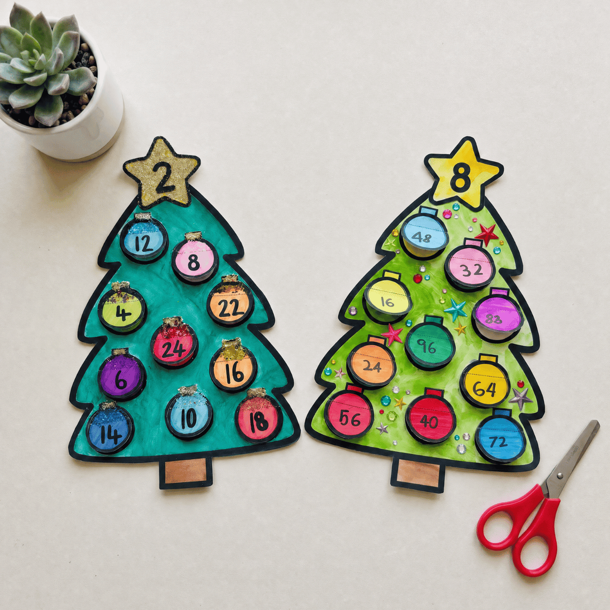 Two completed Christmas tree multiplication craftivities, a teal tree with 2 on the star and a green tree with 8 on the star, each decorated with ten coloured baubles showing the answers to that times table, beside a pair of red scissors.