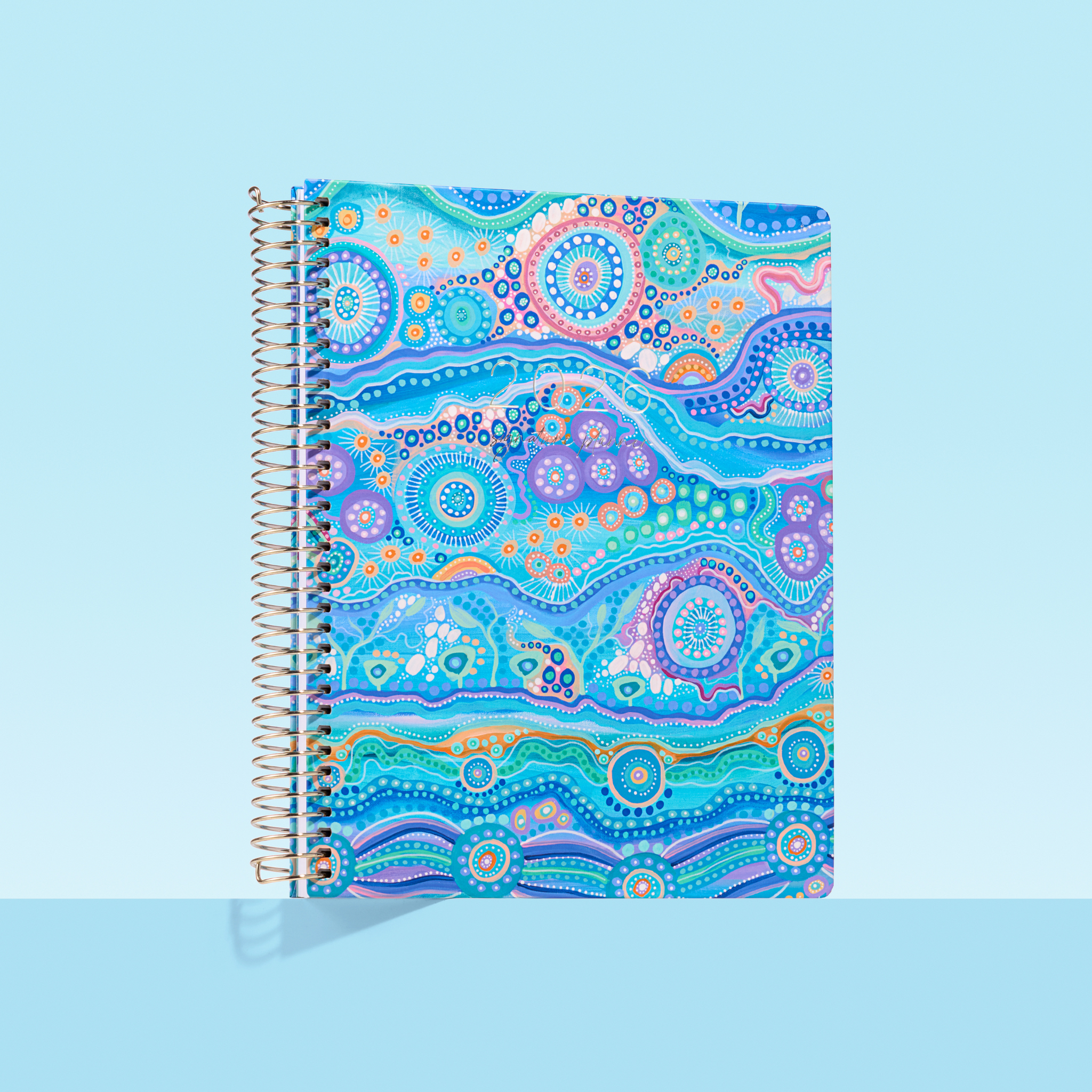 Blue 2026 spiral-bound planner featuring Aboriginal-inspired artwork with flowing coastal patterns, circles, and dots in ocean tones on a light blue background.