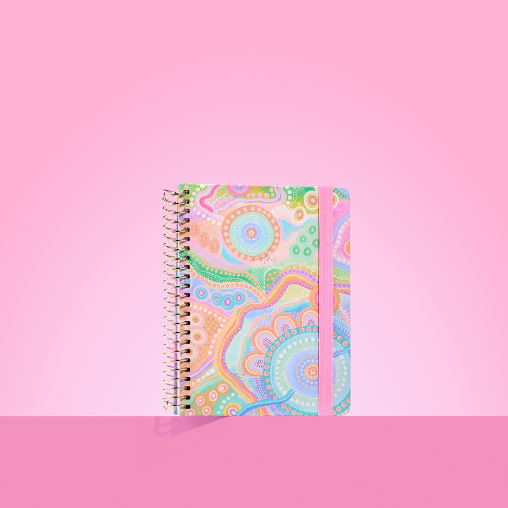 Pastel-toned 2026 notepad with Aboriginal-inspired circular patterns, gold spiral binding, and pink elastic closure on a pink background.